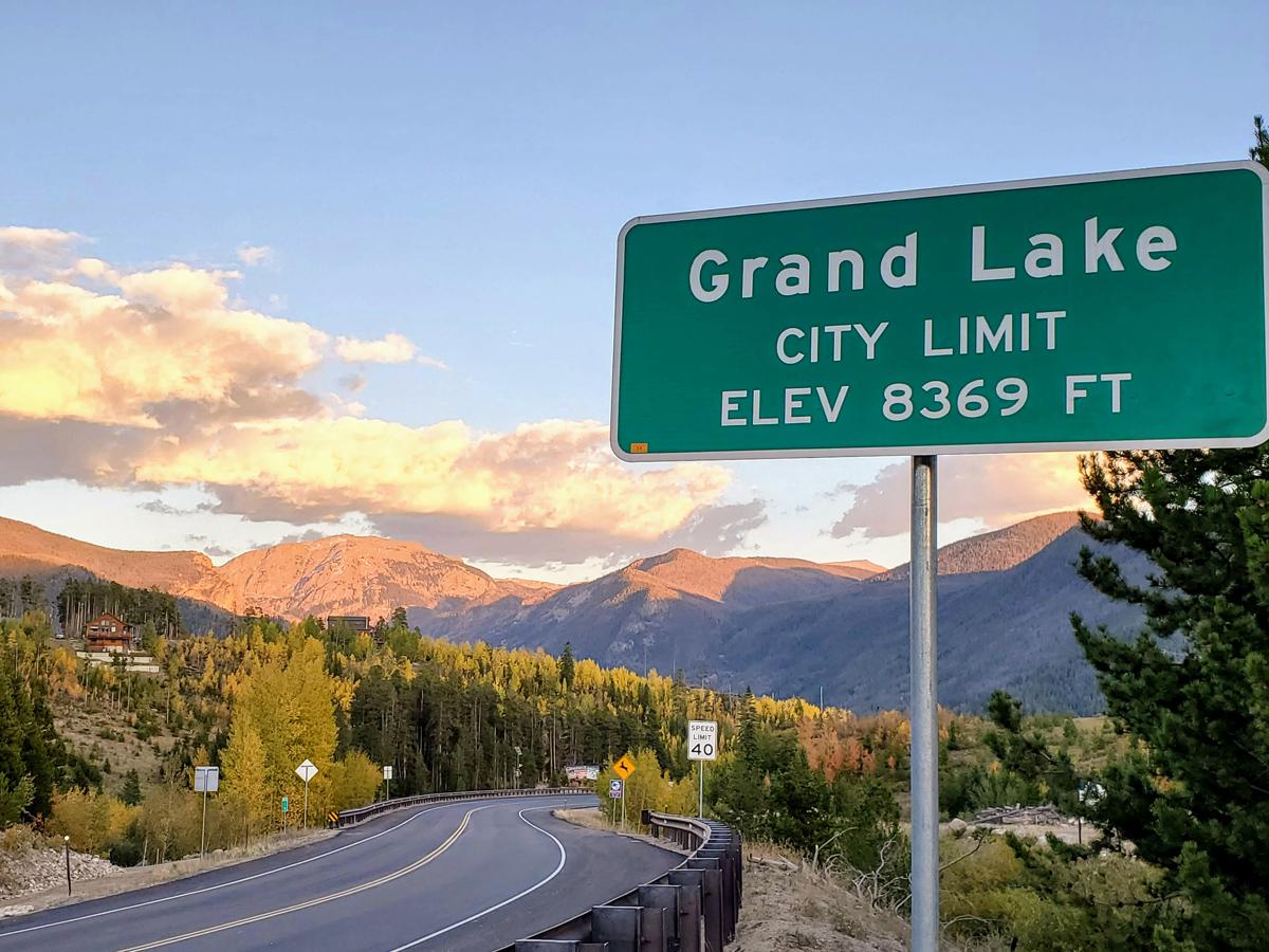 Road curving past a sign that says Grand Lake and lists an elevation of 8,369 feet with evergreens and mountains in the background.