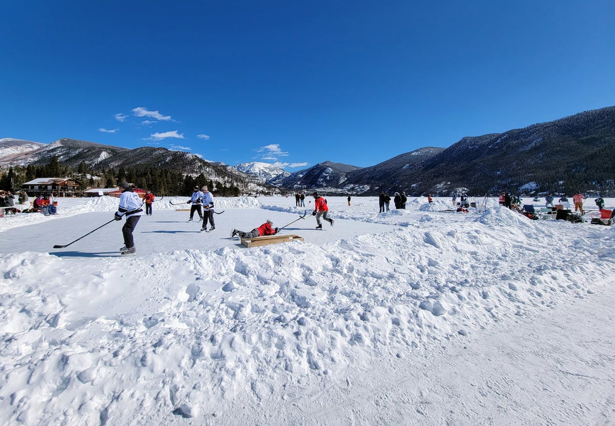 People holding hockey sticks practice their shots during the Pond Hockey Classic. There's a blue sky and mountains in the distance.