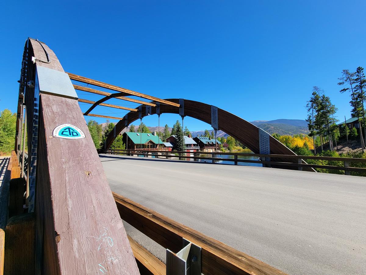 A metal, arched road bridge with a bright-blue sky sits on an early fall day. There are evergreen trees and trees beginning to turn golden.