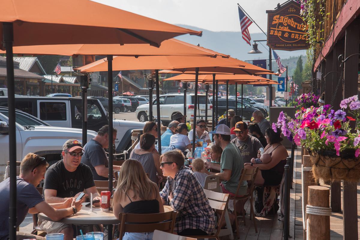 People eat at outdoor cafe tables with opened, orange umbrellas shading them from the sun at Sagebrush BBQ & Grill along the Grand Lake Boardwalk.