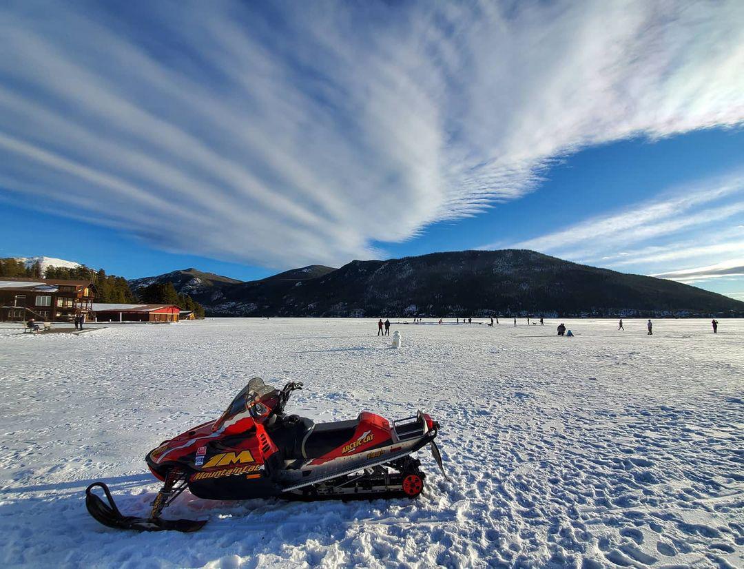 A red snowmobile sits on the frozen Grand Lake. In the background there are people walking around. In the distance a mountain rises to meet a blue sky with white clouds.