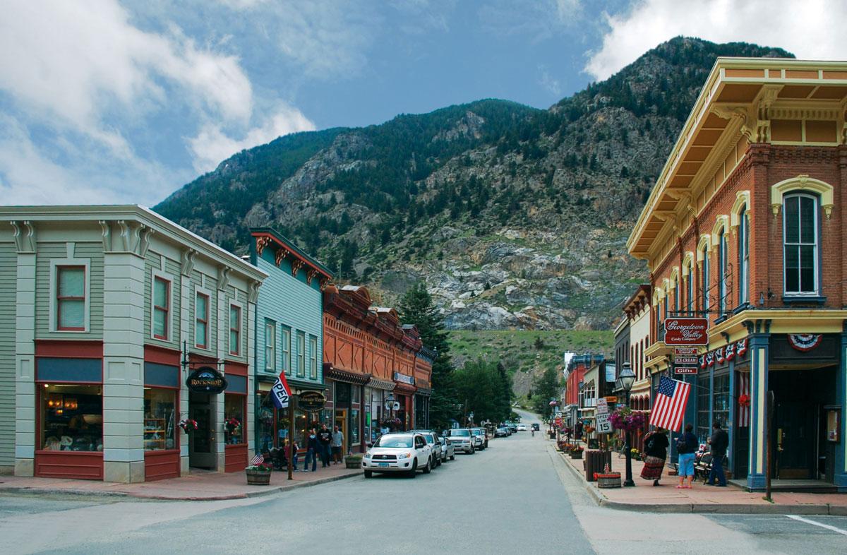 A view of a Georgetown street in the summer with a mountain in the distance meeting a blue sky with a couple clouds. The buildings that line the street are Victorian, some are painted pastel colors, others are red-brick.