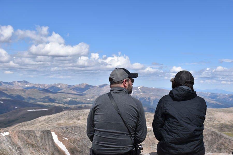 Two people sit at the 14,265-foot summit of Mount Blue Sky looking at other mountains in the distance