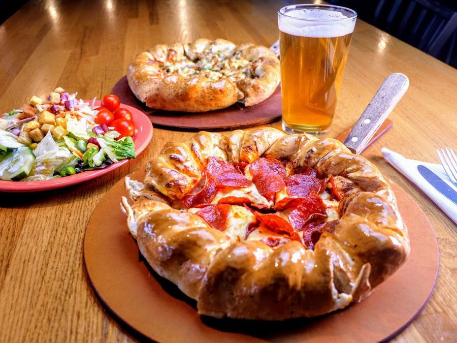 A pepperoni pizza with a very thick crust pictured with a beer a salad and another pizza in the background