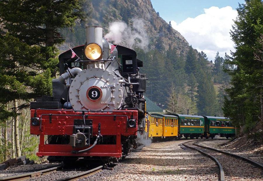 On a summer's day, surrounded by a mountain and evergreen trees the engine that's black with a red bottom leads yellow and green train cars of the Georgetown Loop Rail Road.