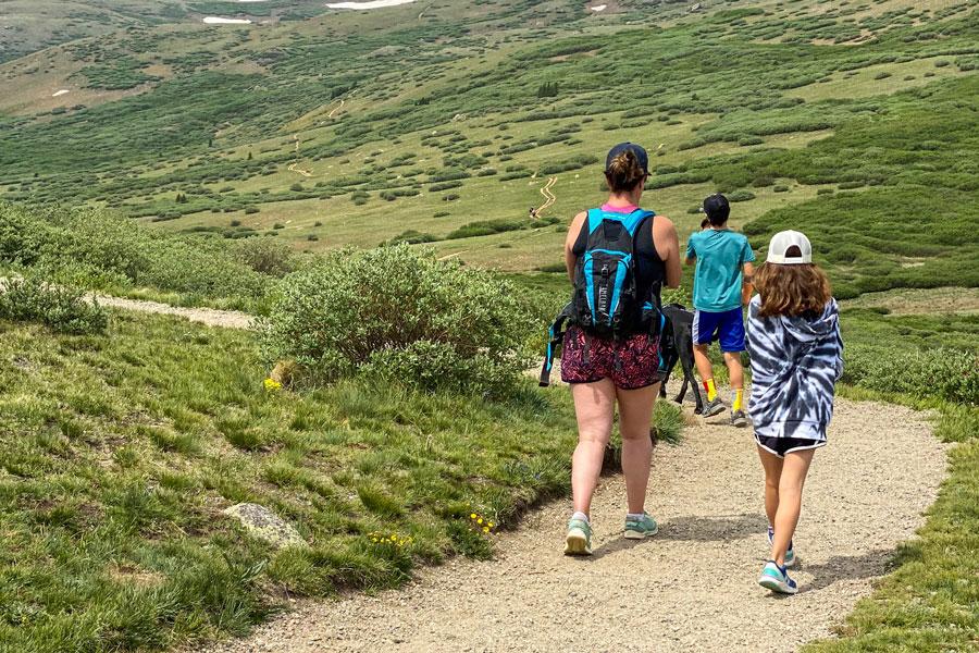 Three people hike on a dirt path surrounded by green grass hills on both sides. One person on the left is wearing a blue backpack with a black top, the person on the left is wearing a tie-dye sweatshirt and in front of them a person in a teal t-shirt walks a black dog.