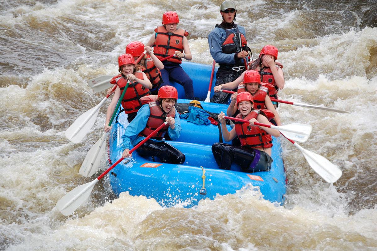 Eight people wearing helmets paddle in a blue raft through whitewater on the Colorado River