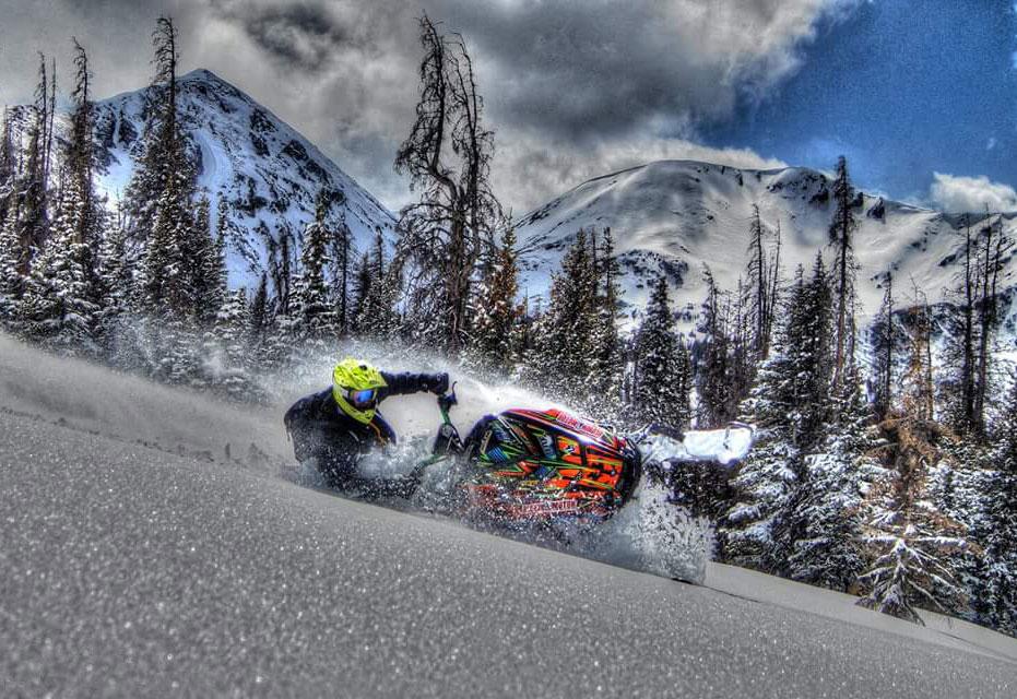 A snowmobiler takes a hard turn with snow-covered peaks and evergreen trees in the background on Gore Pass