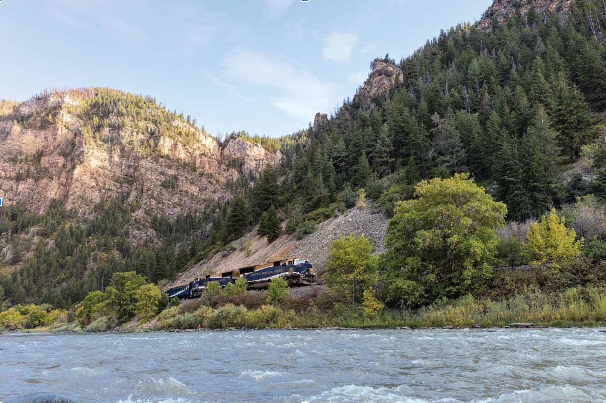 The Rocky Mountaineer train travels alongside the rushing Colorado River as the sun sets behind a rocky outcrop in Colorado.