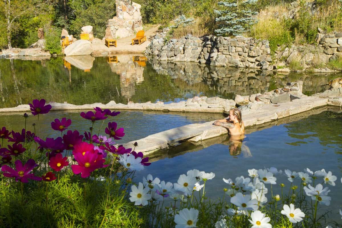 A woman rests on the edge of the Strawberry Park Hot Springs pool on a sunny day, with pink and white wildflowers blooming in the foreground