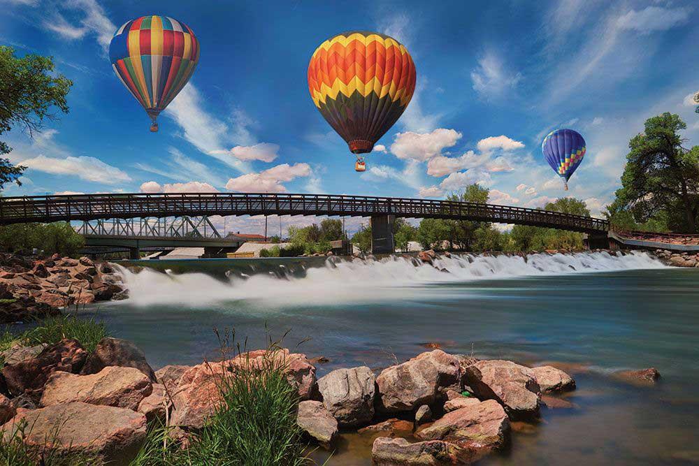 A rainbow and brown, red and orange hot-air balloon sail above a bridge over the rushing rapids of the Arkansas River in Pueblo