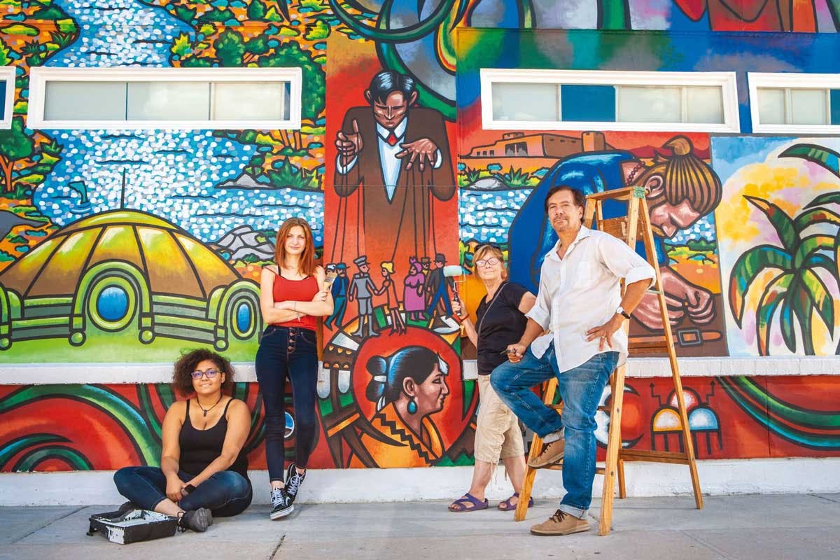 Four artists stand in front of a vibrant mural mural along Pueblo's Creative Corridor