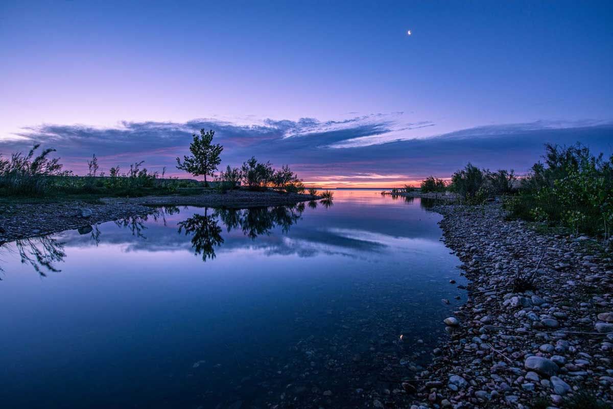 Sun sets beneath a lavendar-colored sky that's reflected in the still wateres of Lake Pueblo along a rocky shore, with litte trees popping up here and there