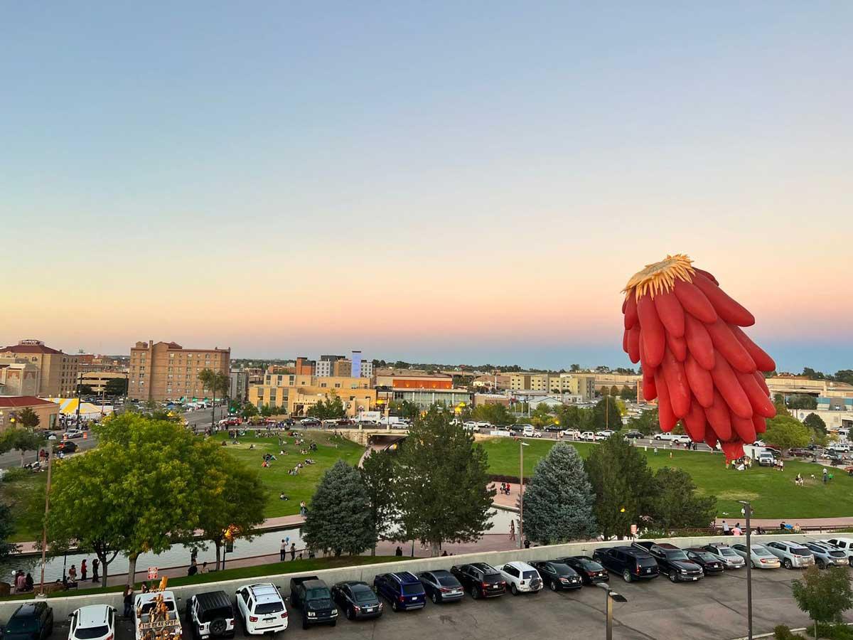 A giant red-chili-shaped balloon hovers over a parkinglot with a view of downtown Pueblo