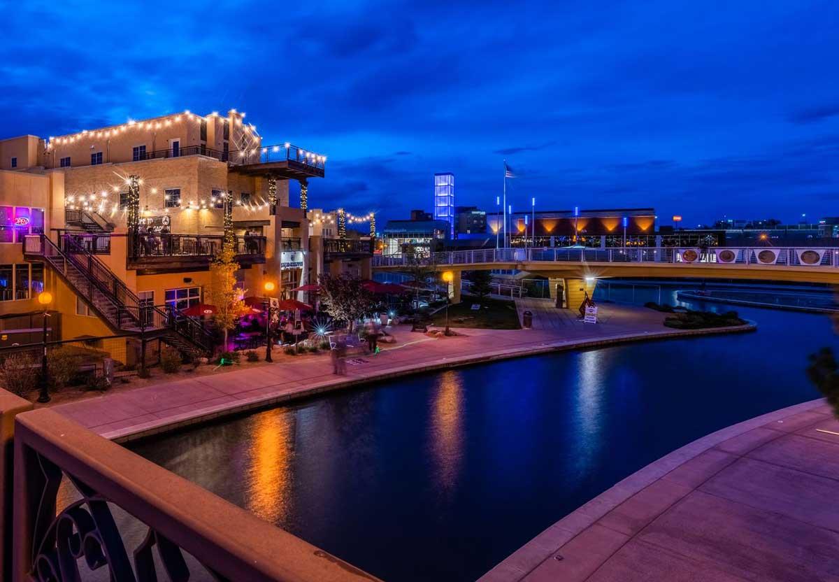 Under a darkening sky, the gently flowing waters of the Arkansas River flow by a restaurant with white lights strung on its balcony