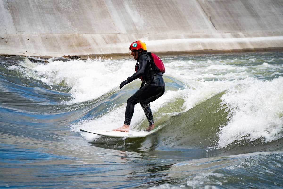 A surfboarder in a black wesuit and red helmet rides waist-high waves between the concret walls of the Pueblo Levee