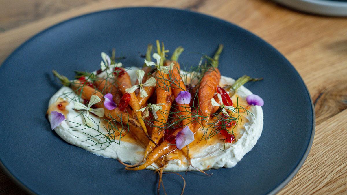 A blue plate holds mini carrots topped with purple flowers on a creamy sauce