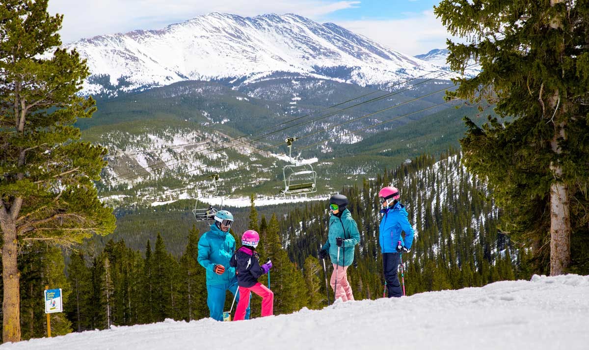 A family of skiers in bright clothing pause on the ski mountain with snowy trees around then and a snowy peak in the distance