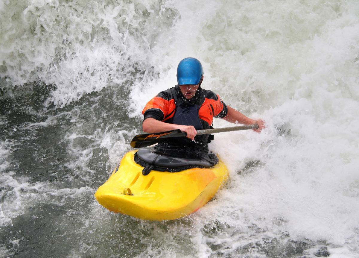 A person wearing a life vest and helmet paddles over a wave in a yellow kayak at the Cache la Poudre Whitewater Park in Fort Collins, Colorado.