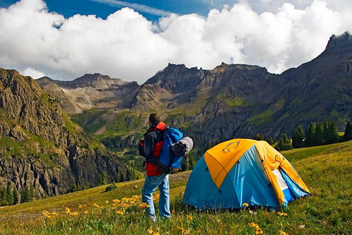 A hiker with a backpack stands next to his tent, facing a mountain ridge