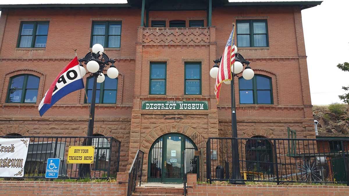 A red-brick historical building in Cripple Creek, Colorado, houses the district museum. An 'OPEN" flag and an American flag waves in the wind outside the building. 