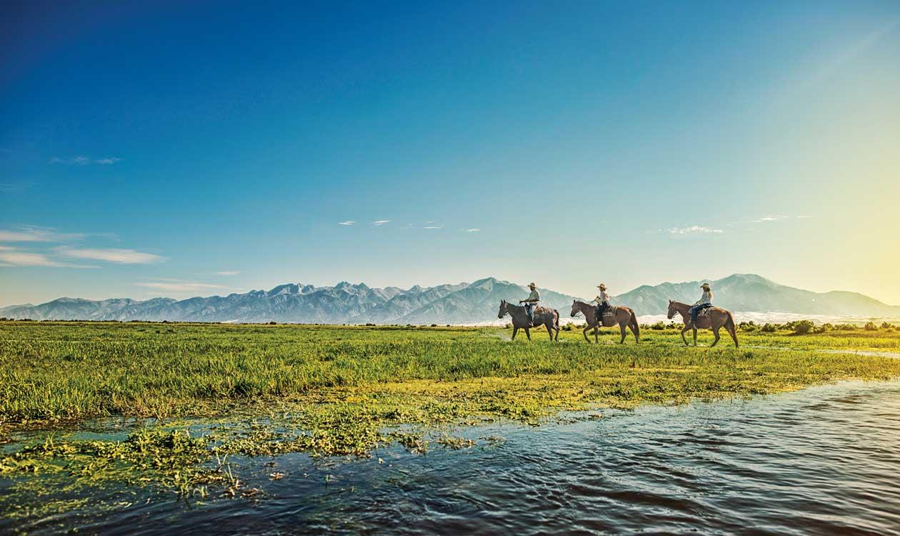 Three people on horseback ride alongside a river