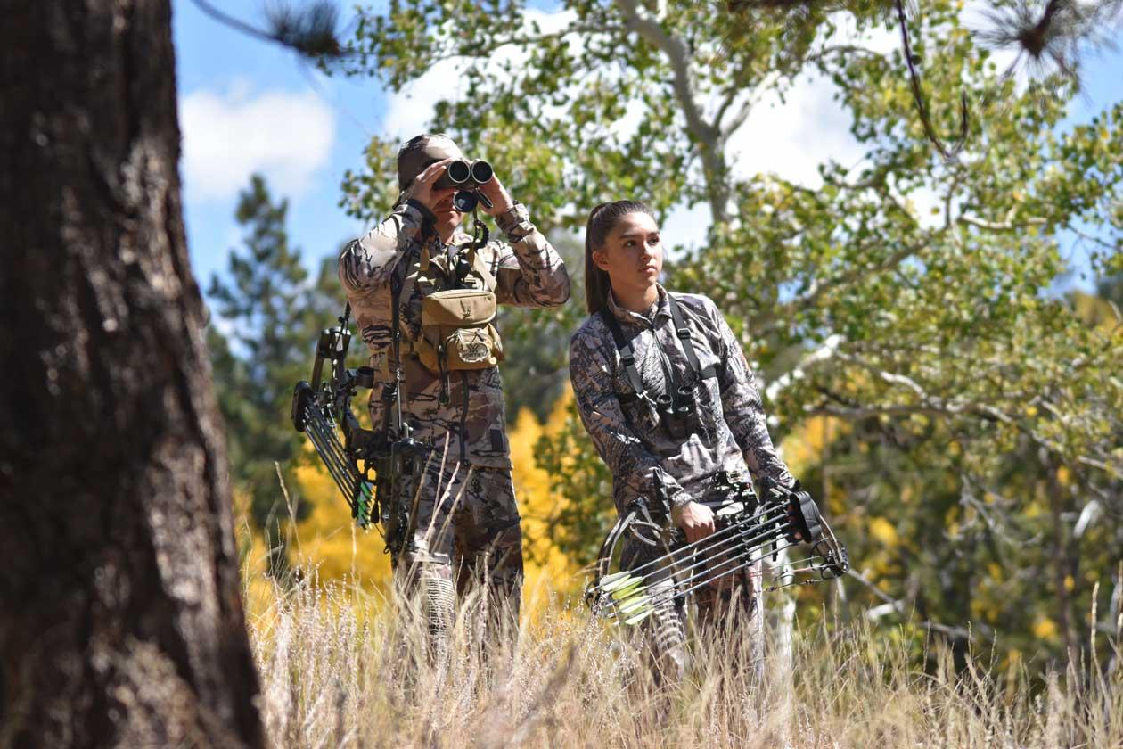 A man and woman in camo carrying bows and arrows look through binoculars