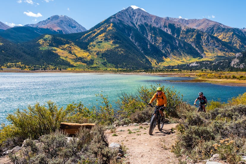 Two people ride mountain bikes around the bright blue waters of Twin Lakes with mountains rising behind them near Leadville, Colorado