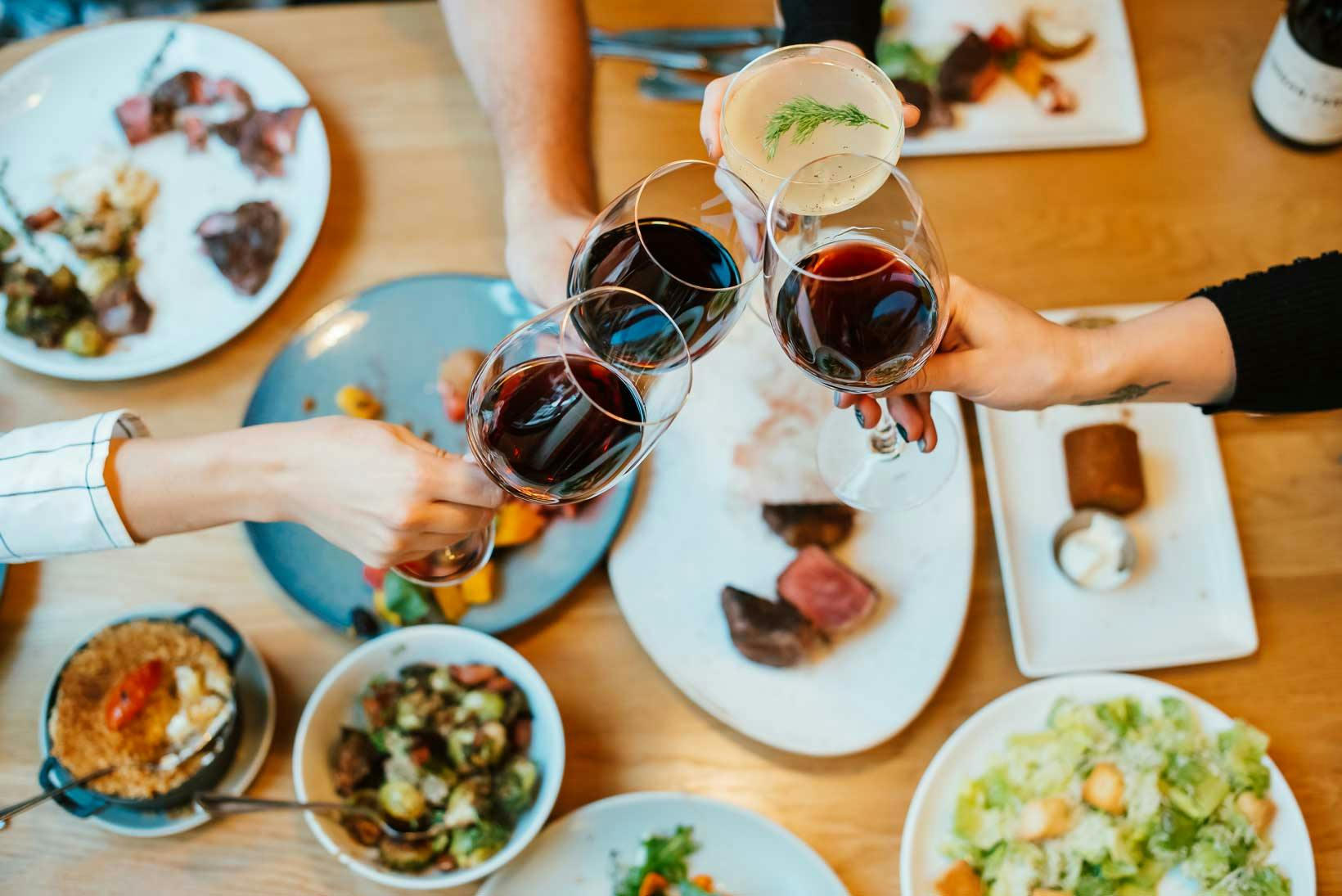 Four people cheers red wine glasses over a table full of plates and food