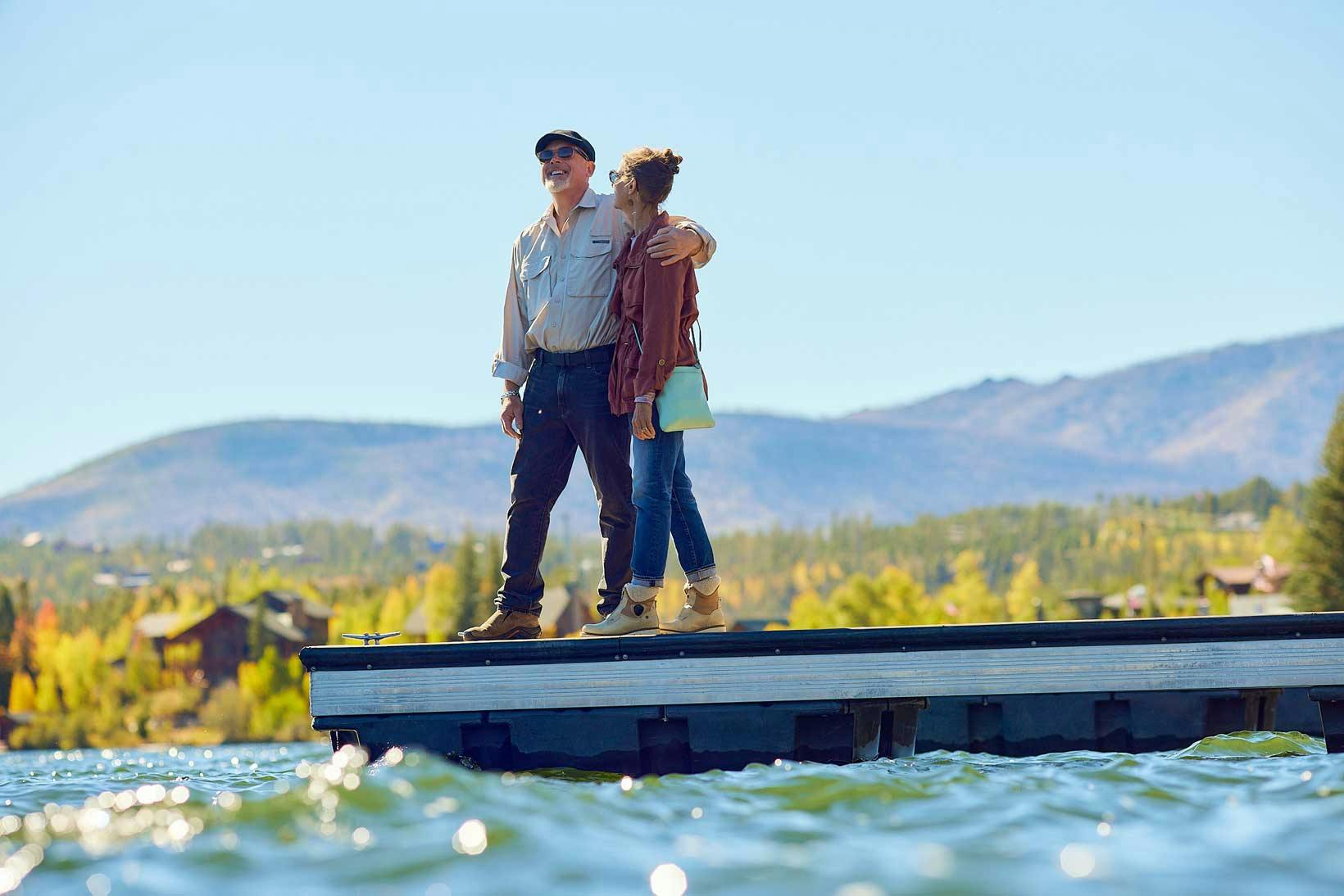 Two people embrace on a boat doc with mountains behind them