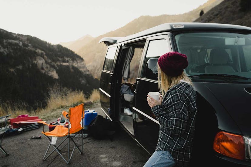 A woman leans on a van and drinks a cup of cofee while admiring mountain views