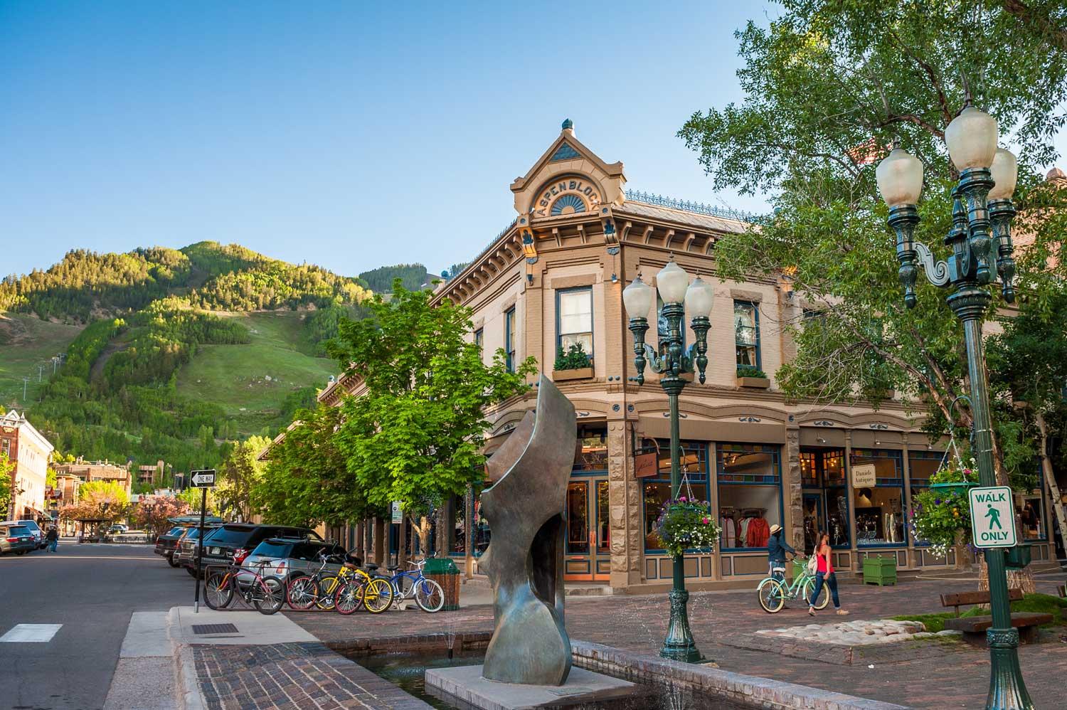 Summer in downtown Aspen. There is a blue sky, the trees have bright-green leaves on them and a historic gold-rush era building has a public art sculpture in front of it with people walking and biking.