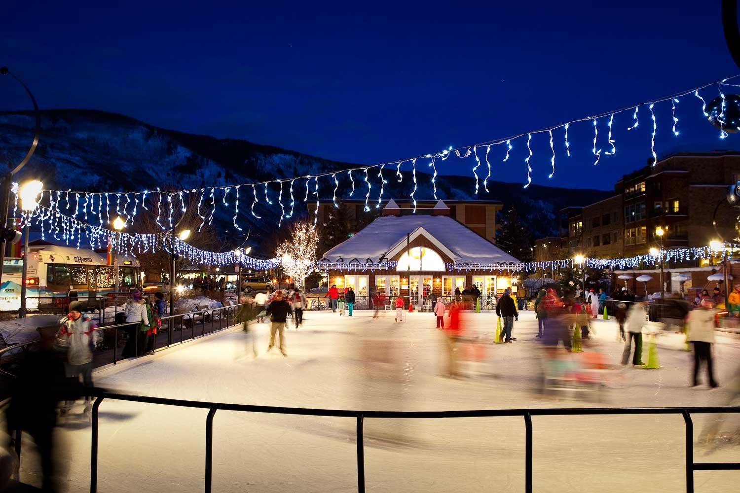 At night, ice skaters make their way around a crowded rink with twinkle lights above the ice.