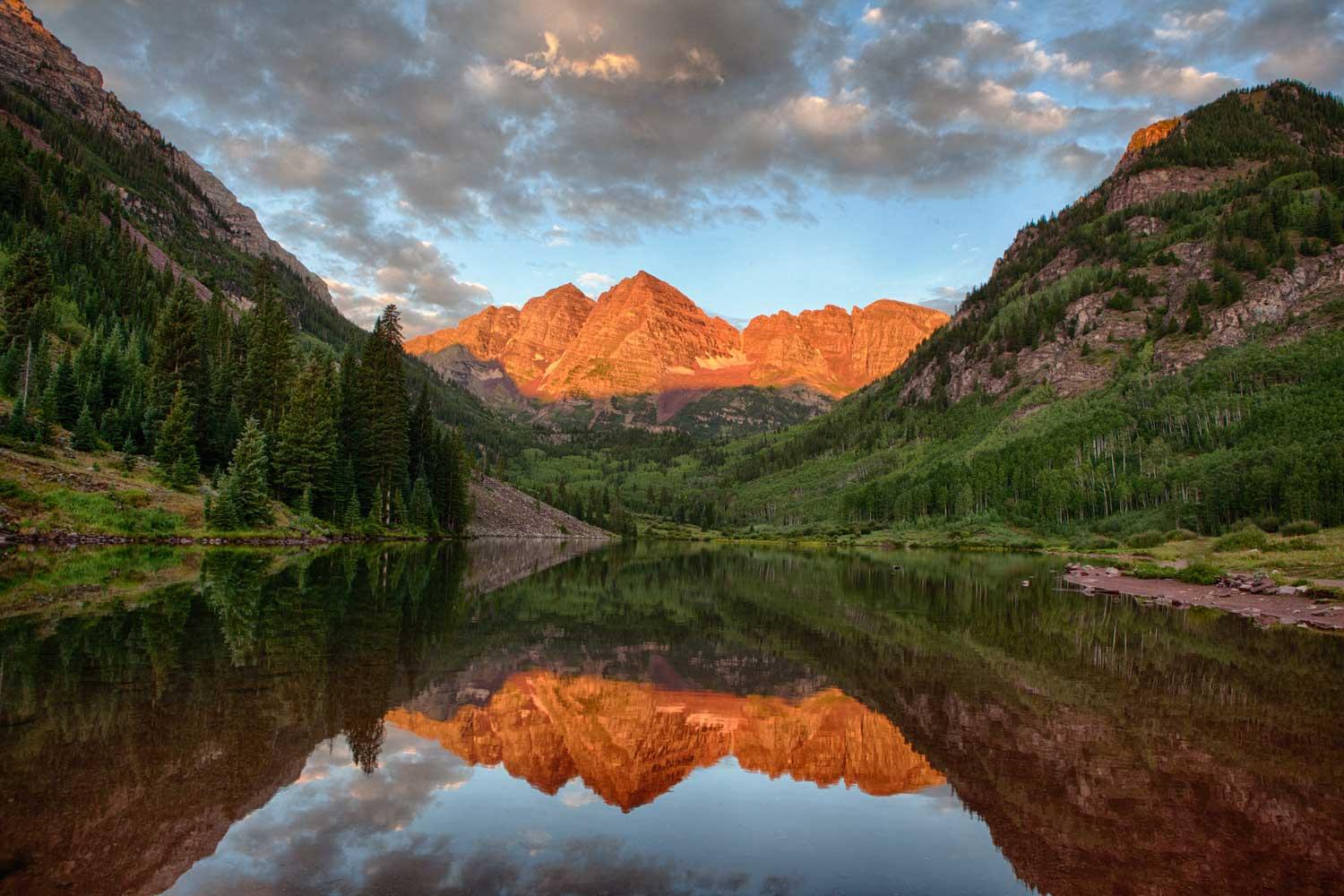 A serene mountain lake with a rocky mountain lit up in a sunset's glow in the background. The sky is blue, the lake is smooth and the surrounding mountain peaks are green and brown.