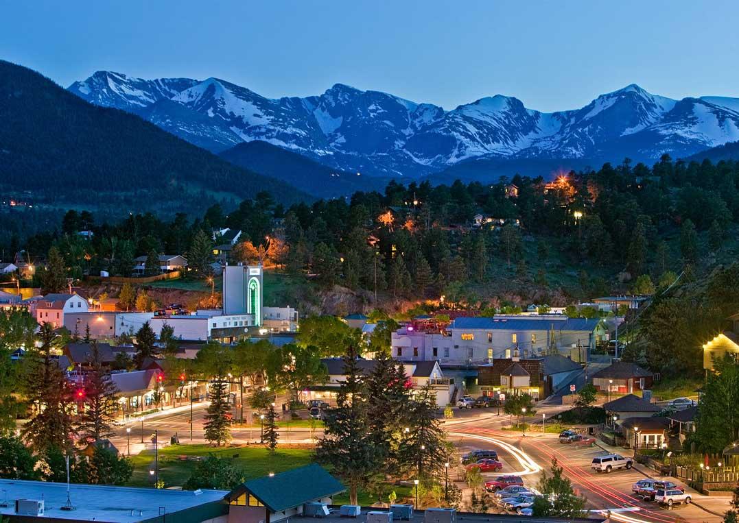 At twilight the town of Estes Park from above. The buildings are lit up and in the distance snow-capped mountains meet a light-blue sky. The trees that surround the town are green.