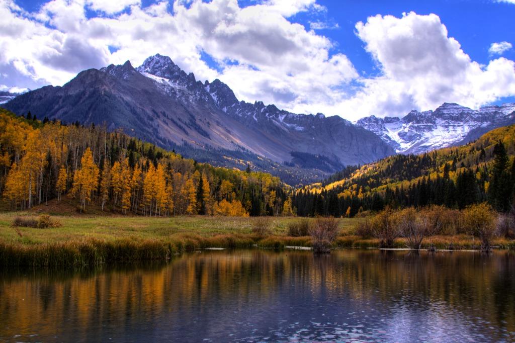 Mount Sneffels' peak is covered in snow in the distance behind a lake with golden aspens and evergreen trees surrounding the lakeshore.