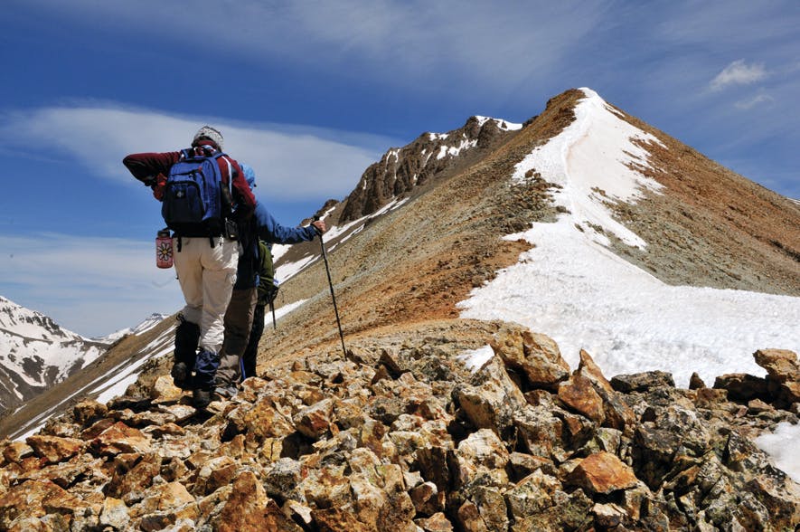 A mountain climber stands on rocky ground and stares up at a mountain peak that's covered in snow. There's a blue sky with slight white clouds. 