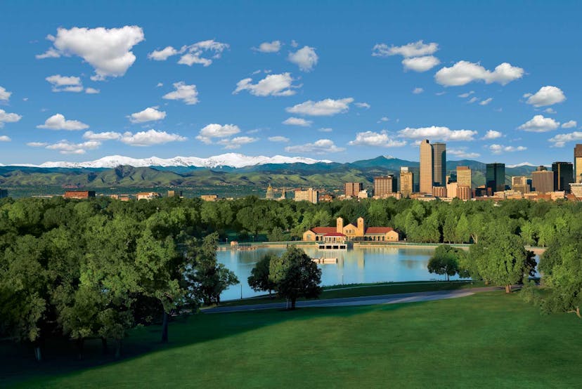 The Denver skyline sits among blue sky mottled with clouds. To the back of the city is the Front Range. In front of the city is a bakehouse, a reflective blue lake and green grass and green trees.
