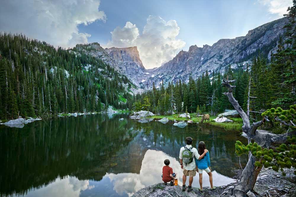 A family of three stands on the edge of a mountain lake looking out at it's glassy surface. There are evergreen trees surrounding the water and the shores have rocks and boulders. In the distance Rocky Mountain peaks soar to meet a light blue sky with clouds.