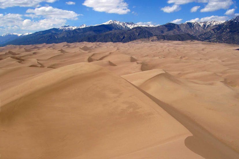 Group of sand dunes at Great Sand Dunes National Park. The sky is blue with the sun shining and there are white clouds. In the background are snow-capped towering mountains.