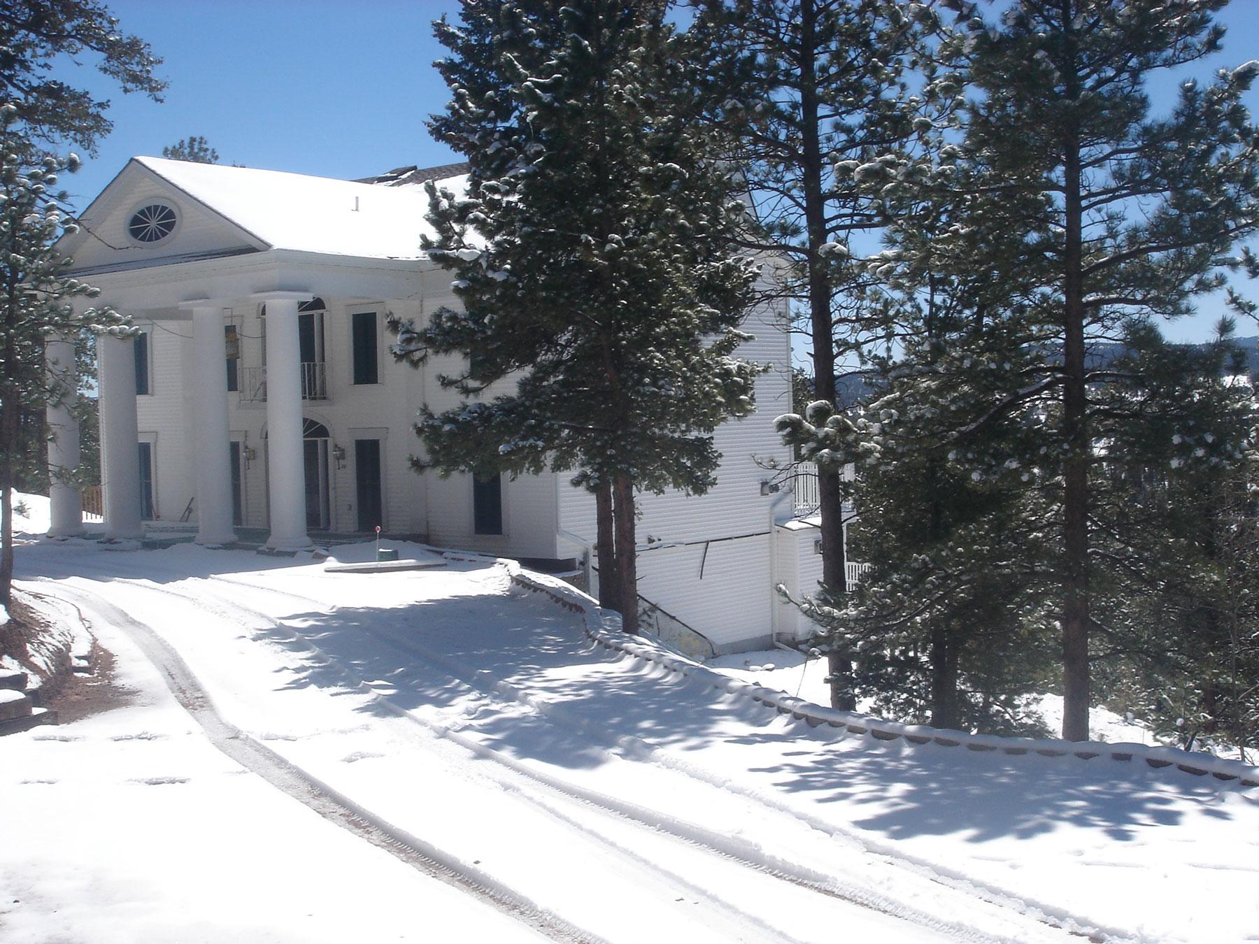 A white house with a pediment and four columns sits in a snow-covered area with evergreen trees. 