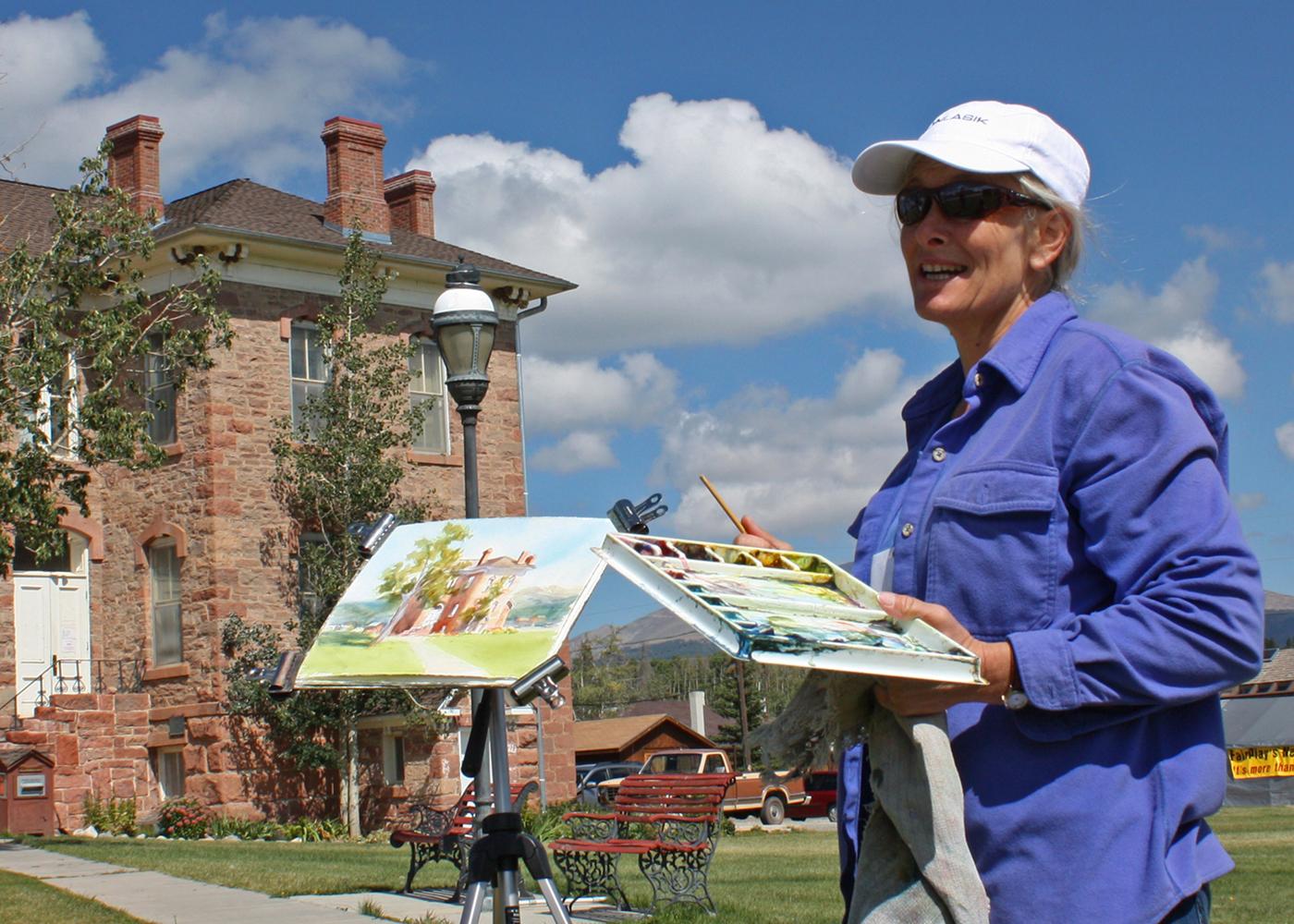 A person in a purple shirt holds a watercolor set and painting on a summer's day at the Art Festival in FairPlay. In the background a historic, two-story red-stone building sits under a blue sky with white clouds. There are a couple green-leafed trees behind the painter.