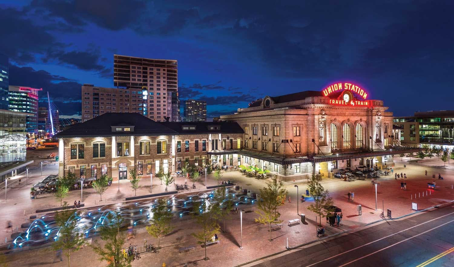 Denver's Union Station at night. The historic building's red neon sign reads "Union Station" above a clock and below the clock it says "Travel By Train." There are small green trees surrounding a square with a water feature. There are people mingling around the station. And the blue night sky features some high rise buildings behind the building.