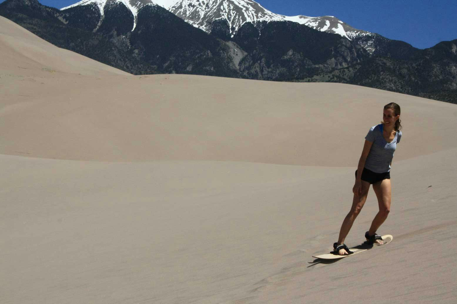 Woman sand boards on a sand dune in Great Sand Dunes National Park with snow-capped mountains in the background. The sky is blue. 