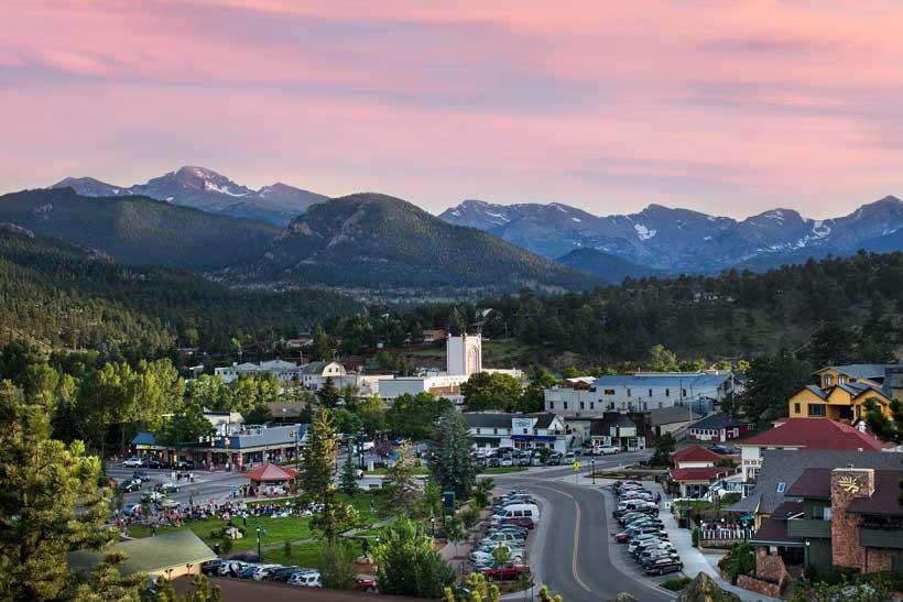 On a summer evening, an aerial view of Estes Park. The trees are green, the sky is a light pink and the Rocky Mountains soar in the distance.