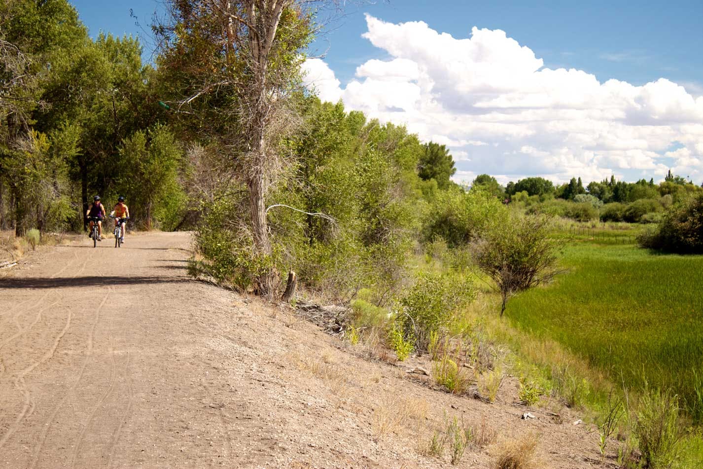 In the left of the image on a dirt trail, two bicyclists are cycling toward the camera in the distance. On the right, a green valley with trees, a blue sky with lots of white cloud coverage. 