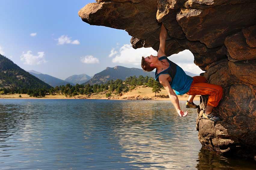 A person boulders in a blue tank top and orange pants on a rock formation at the edge of Marys Lake. In the distance evergreen trees sit on a yellow lakeshore with a blue sky and white clouds and mountains.