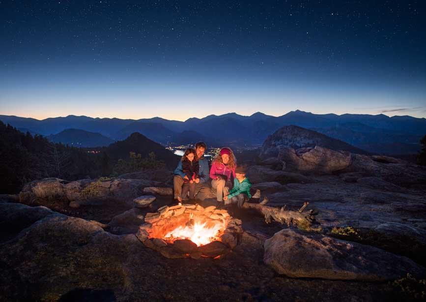 A family at night sits in front of a fire. The fire pit sits among boulders and in the distance mountain peaks reach for a light blue sky that turns black.