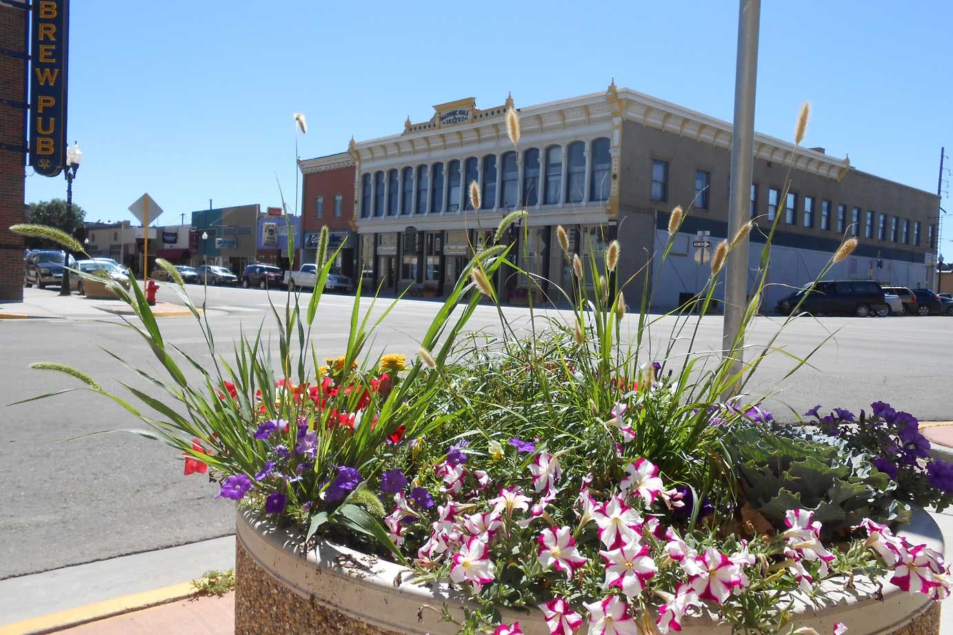 Downtown Alamosa is in the background with a planter of spring flowers in the foreground. It’s a blue sky day with a historic Masonic aHall in the background. There are lots of cars parked on the street. 