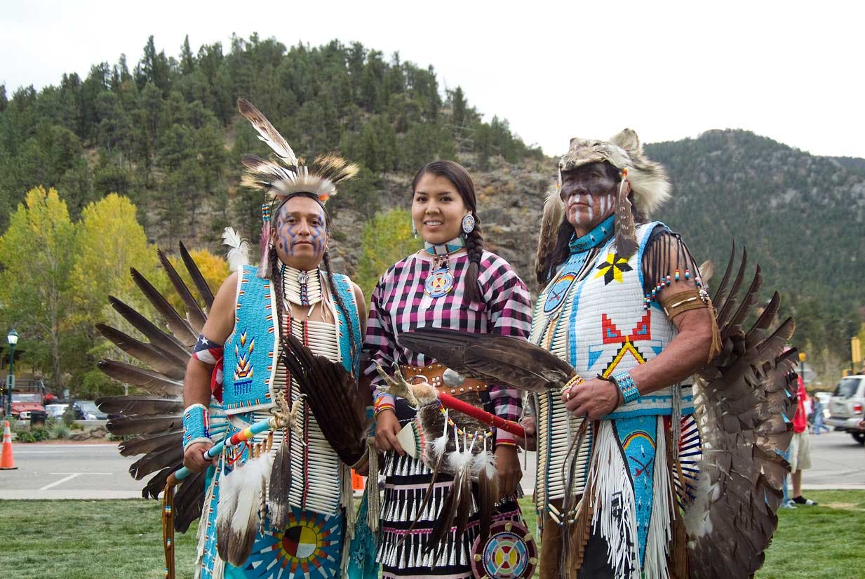 Three American Indian dancers pose on a summer's day at the Estes Park Elk Festival. In the background evergreen trees cover hills.